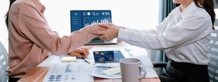 Two Asian businesswomen shaking hands in modern office after successfully analyzing piles of...
