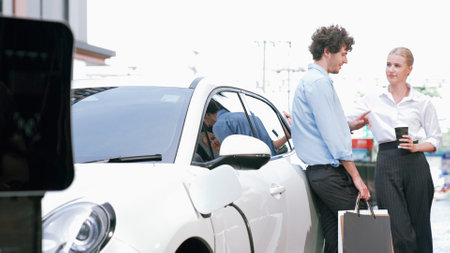 Progressive businessman and businesswoman leaning on electric car connected to charging station before driving around city center. Eco friendly rechargeable car powered by clean energy.の写真素材