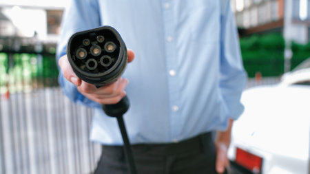 Suit-clad businessman with progressive ambition leaning pointing power cable plug at camera, charging station at city center. Electric vehicle, EV car driven by renewable energy for green environment.の写真素材