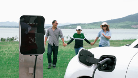 Concept of progressive happy family enjoying their time at wind farm with electric vehicle. Electric vehicle driven by clean renewable energy from wind turbine generator for charging station.の写真素材