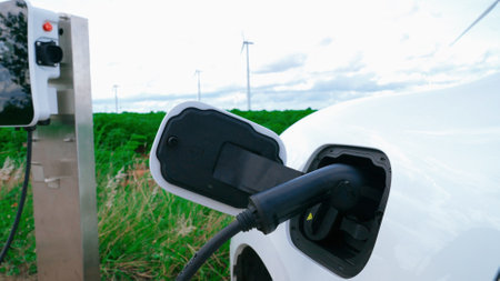 Progressive man with his electric car, EV car recharging energy from charging station on green field with wind turbine as concept of future sustainable energy. Electric vehicle with energy generator.の写真素材