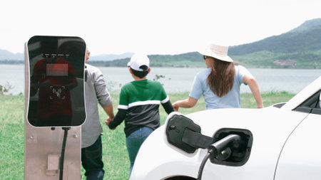 Concept of progressive happy family enjoying their time at wind farm with electric vehicle. Electric vehicle driven by clean renewable energy from wind turbine generator for charging station.の写真素材