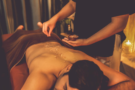Man customer having exfoliation treatment in luxury spa salon with warmth candle light ambient. Salt scrub beauty treatment in Health spa body scrub. Quiescentの写真素材