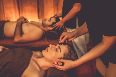 Caucasian couple enjoying relaxing anti-stress head massage with hot stone and pampering facial beauty skin recreation leisure in warm candle light ambient salon spa in resort or hotel. Quiescentの写真素材