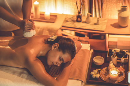 Hot herbal ball spa massage body treatment, masseur gently compresses herb bag on woman body. Tranquil and serenity of aromatherapy recreation in warm lighting of candles at spa salon. Quiescentの写真素材