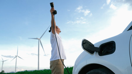 Progressive man with his electric car, EV car recharging energy from charging station on green field with wind turbine as concept of future sustainable energy. Electric vehicle with energy generator.の写真素材