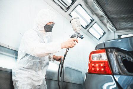 Automotive service worker in full protective gear expertly apply color paint in to cars bodywork with spray gun or respirator painting in chamber workshop. Car paint service for scratch refinish.Oxusの写真素材