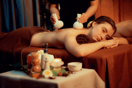Hot herbal ball spa massage body treatment, masseur gently compresses herb bag on couple customer body. Serenity of aromatherapy recreation in warm lighting of candles at spa salon. Quiescentの写真素材