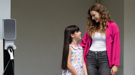 Happy little young girl learn about eco-friendly and energy sustainability as she help her mother recharge electric vehicle from home EV charging station. EV car and modern family. Panorama Synchronosの写真素材