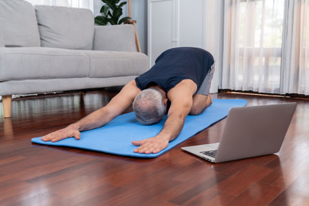 Senior man in sportswear being doing yoga in meditation posture on exercising mat at home. Healthy senior pensioner lifestyle with peaceful mind and serenity. Cloutの写真素材