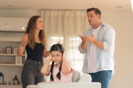 Annoyed and unhappy young girl sitting on sofa trapped in middle of tension by her parent argument in living room. Unhealthy domestic lifestyle and traumatic childhood develop to depression.Synchronosの写真素材