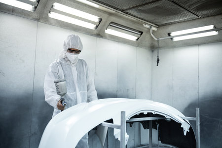 Automotive service worker in full protective gear expertly apply color paint in to cars bodywork...