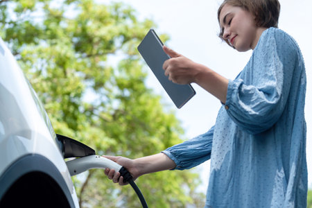 Holiday road trip vacation by the green countryside nature with beautiful young woman checking battery status from tablet while recharging electric vehicle.Eco-friendly travel wit EV car.Perpetualの写真素材