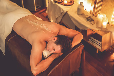 Man customer having exfoliation treatment in luxury spa salon with warmth candle light ambient. Salt scrub beauty treatment in Health spa body scrub. Quiescentの写真素材