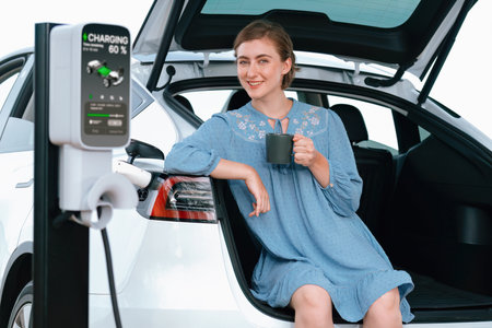 Holiday road trip vacation with young woman sitting in the car trunk with coffee while recharging...