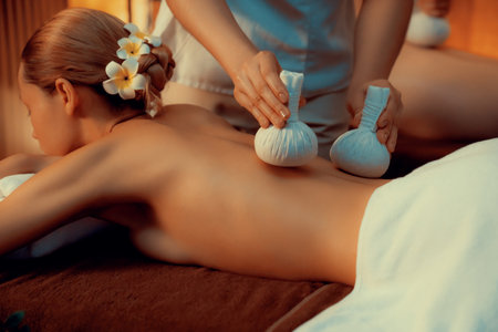 Hot herbal ball spa massage body treatment, masseur gently compresses herb bag on couple customer body. Serenity of aromatherapy recreation in warm lighting of candles at spa salon. Quiescentの写真素材
