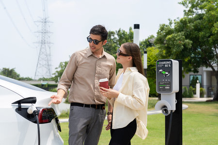 Young couple with coffee pay for electricity while recharge EV car battery at charging station...