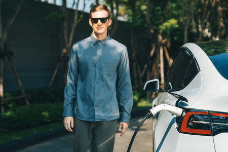 Young man travel with EV electric car charging in green sustainable city outdoor garden in summer shows urban sustainability lifestyle by green clean rechargeable energy of electric vehicle innardsの写真素材