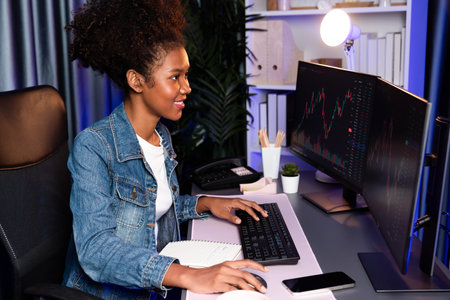 Young African woman trader smiling on happy face, looking on screen with valued stock market chart...