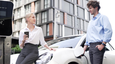 Progressive businessman and businesswoman leaning on electric car connected to charging station...