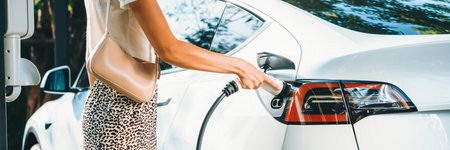 Young woman travel with EV electric car charging in green sustainable city outdoor garden in summer....
