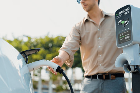 Young man put EV charger to recharge electric cars battery from charging station in city commercial parking lot. Rechargeable EV car for sustainable environmental friendly urban travel. Expedientの写真素材