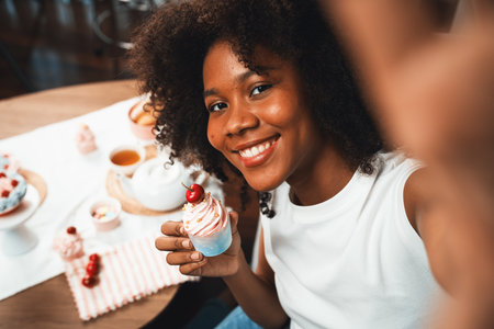Beautiful young African blogger presenting piece of cupcake in concept special cuisine with selfie on smartphone. Content creating of social media with favorite sweets bakery dish. Tastemaker.の写真素材