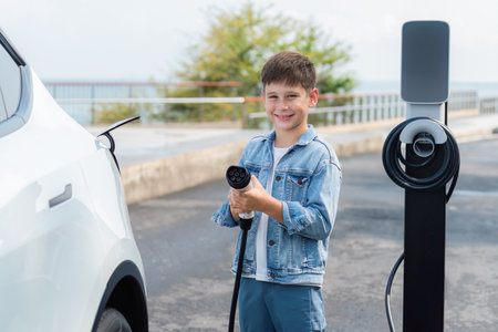 Playful little boy pointing EV charger at camera, recharging eco-friendly electric car from EV charging station. EV car travel by the seashore using clean and sustainable energy.Perpetualの写真素材