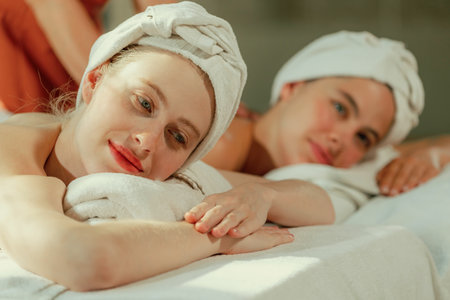 A couple of beautiful young woman with white towel lie on comfortable spa bed during receive relaxing body massage by professional masseur surrounded at spa salon. Gray background. Tranquility.の写真素材
