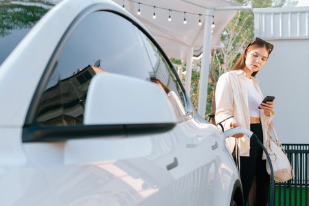 Young woman use smartphone to pay for electricity at public EV car charging station green city park. Modern environmental and sustainable urban lifestyle with EV vehicle. Expedientの写真素材