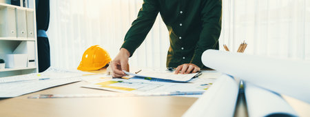 A portrait of professional male engineer shows mistake point in blueprint on messy table with...