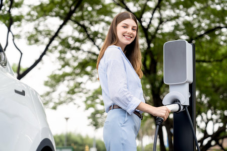Young woman recharge EV electric vehicles battery from EV charging station in outdoor green city park scenic. Eco friendly urban transport and commute with eco friendly EV car travel. Exaltの写真素材