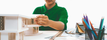 A cropped imaged of female designer measures a house model with a pencil, focusing intently on her...