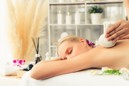 Hot herbal ball spa massage body treatment, masseur gently compresses herb bag on woman body. Tranquil and serenity of aromatherapy recreation in day lighting ambient at spa salon. Quiescentの写真素材