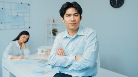 Professional asian businessman crosses arms with confident while young beautiful caucasian colleague drawing a blueprint on meeting table with house model and blueprint at modern office. Immaculate.の写真素材
