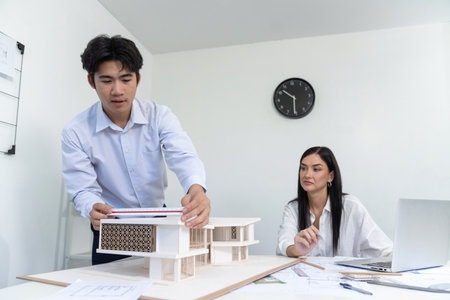 Professional male asian architect using ruler to measure house model length while young beautiful caucasian colleague using laptop to analyzed data on meeting table with house model. Immaculate.の写真素材