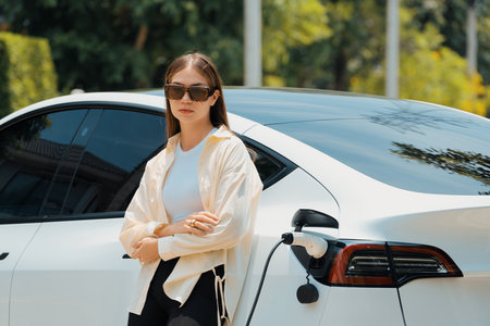 Young woman recharge her EV electric vehicle at green city park parking lot. Urban sustainability lifestyle for environmental friendly EV car with battery charging station. Expedientの写真素材