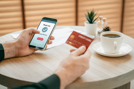 Young man sitting at table using online payment app and digital wallet on smartphone to pay with credit card. E commerce shopping and modern purchasing via mobile internet. Unveilingの写真素材