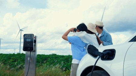 Concept of progressive happy family enjoying their time at wind farm with electric vehicle. Electric vehicle driven by clean renewable energy from wind turbine generator for charging station.の写真素材