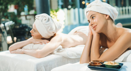 A couple of beautiful young woman relaxes on a spa bed surrounded by nature at outdoor. ready for a body massage. Attractive females in white towels lie on bed peacefully. Close up. Tranquilityの写真素材