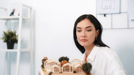 Close up of skilled young beautiful caucasian architect lift house model to check and focus on inspecting mistake point at modern office with blueprint. Creative design and living concept. Immaculate.の写真素材