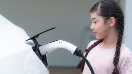 Happy little young girl learn about eco-friendly and energy sustainability as she recharge electric vehicle from home EV charging station. EV car and sustainable future generation concept. Synchronosの写真素材