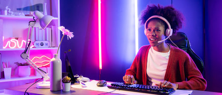 African American girl streamer playing online fighting with Esport skilled team wearing headphones in neon color lighting room. Talking other players planing strategies to win competitors. Tastemaker.の写真素材