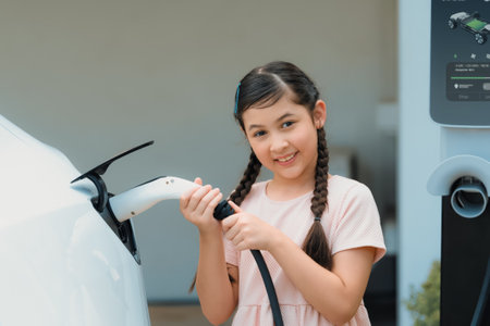 Happy little young girl learn about eco-friendly and energy sustainability as she recharge electric vehicle from home EV charging station. EV car and sustainable future generation concept. Synchronosの写真素材