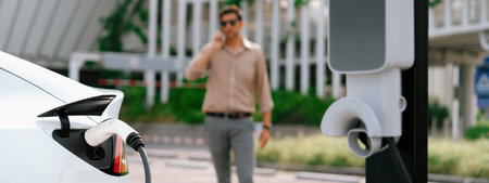 Young man recharge EV electric vehicle at green city mall parking lot while talking on phone. Sustainable urban lifestyle for eco friendly EV car with battery charging station. Panorama Expedientの写真素材