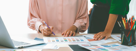 Cropped image of two female young beautiful graphic designer choose the color from color palate by using laptop on table with work tools and equipments scatter around at modern office. Variegated.の写真素材