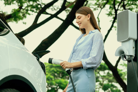Young woman recharging battery for electric car during road trip travel EV car in natural forest or national park. Eco friendly travel during vacation and holiday. Exaltの写真素材