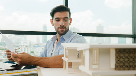 Portrait of smart skilled engineer checking house model while holding blueprint. Professional...
