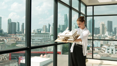 Young smart caucasian beautiful architect engineer raised house model while inspect carefully surrounded by city skyscraper view. Civil engineering, architectural studio. project planing. Traceryの写真素材