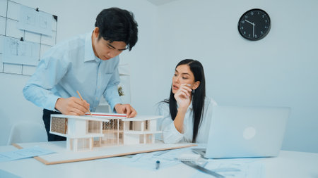Professional male asian architect using ruler to measure house model length while young beautiful caucasian colleague using laptop to analyzed data on meeting table with house model. Immaculate.の写真素材